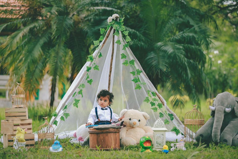 Baby boy sitting in a decorated teepee tent with teddy bears and toys on green grass outdoors.