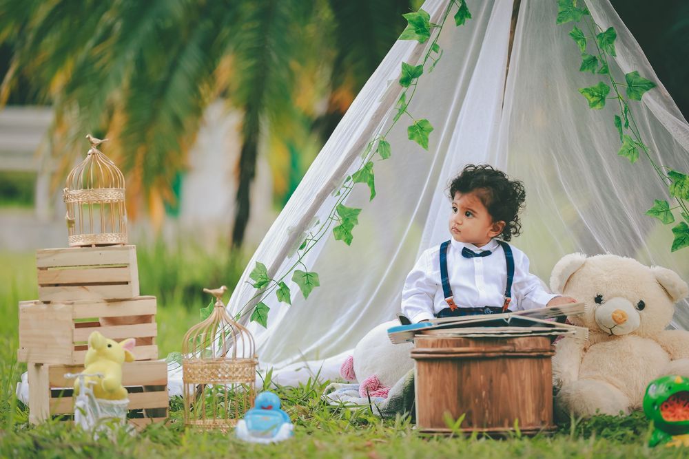 A toddler reads a book beside a white teepee tent with ivy, surrounded by wooden crates, a birdcage, and a large teddy bear.