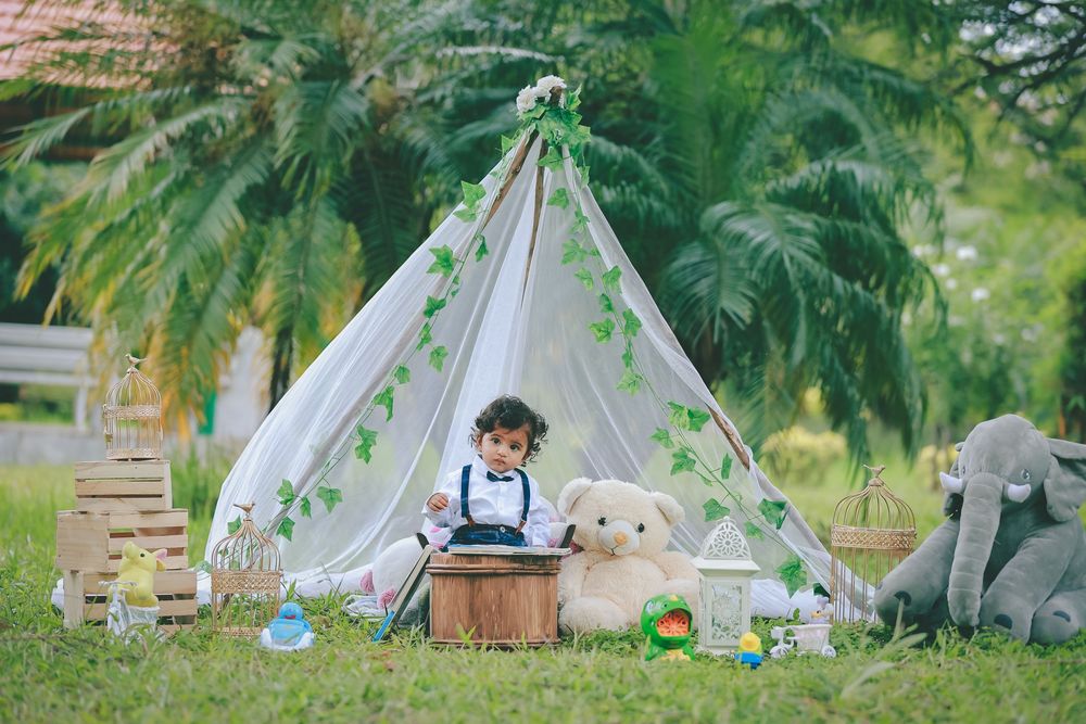 Toddler sitting with teddy bear and elephant toy in a decorated teepee tent outdoors surrounded by lush green trees.
