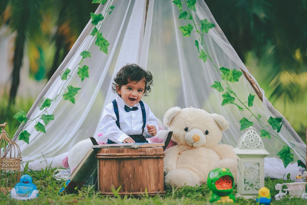 A cheerful toddler in suspenders sits beside a large teddy bear inside a decorated teepee tent outdoors.