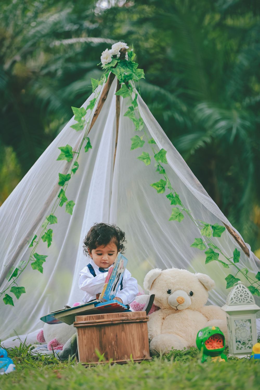 Toddler reading a book beside a teddy bear inside a decorated white teepee tent outdoors.
