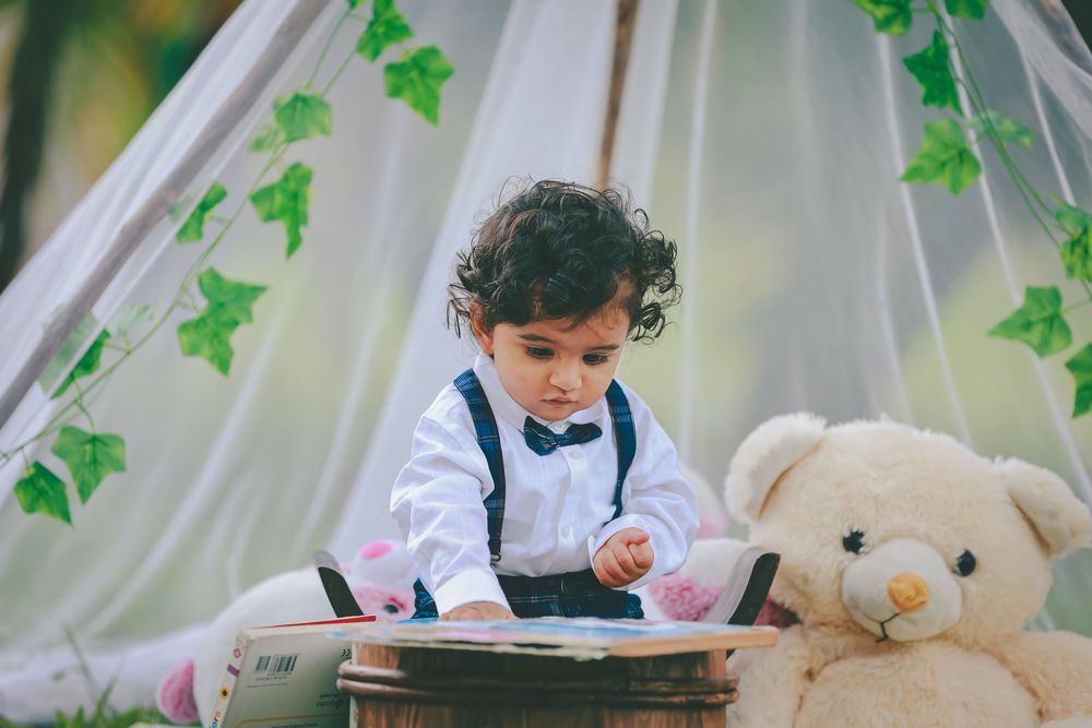 A toddler in suspenders and bow tie reads a book beside a teddy bear under a leafy white tent outdoors.