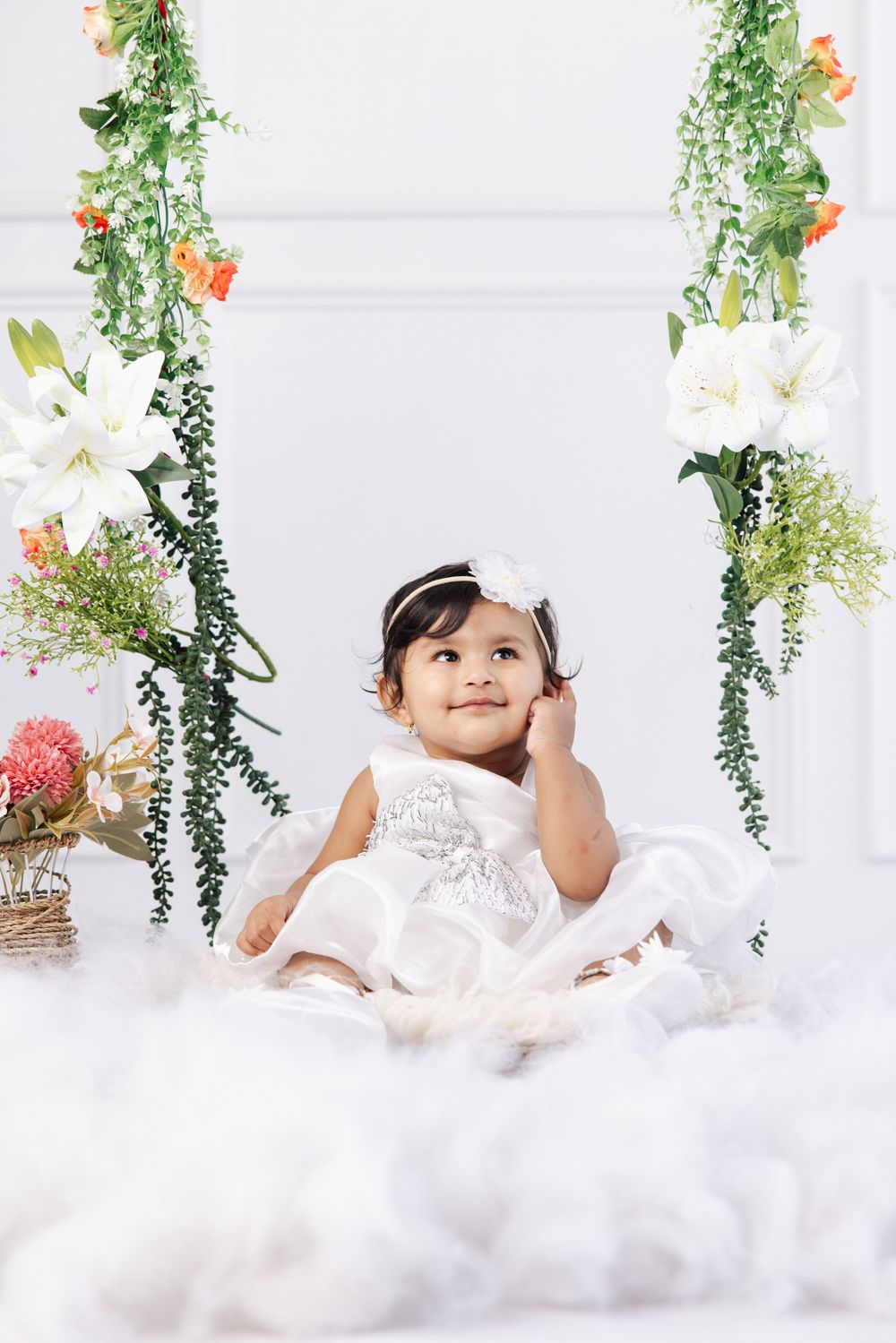 Adorable baby girl in white dress sitting on floral swing surrounded by white flowers and greenery.
