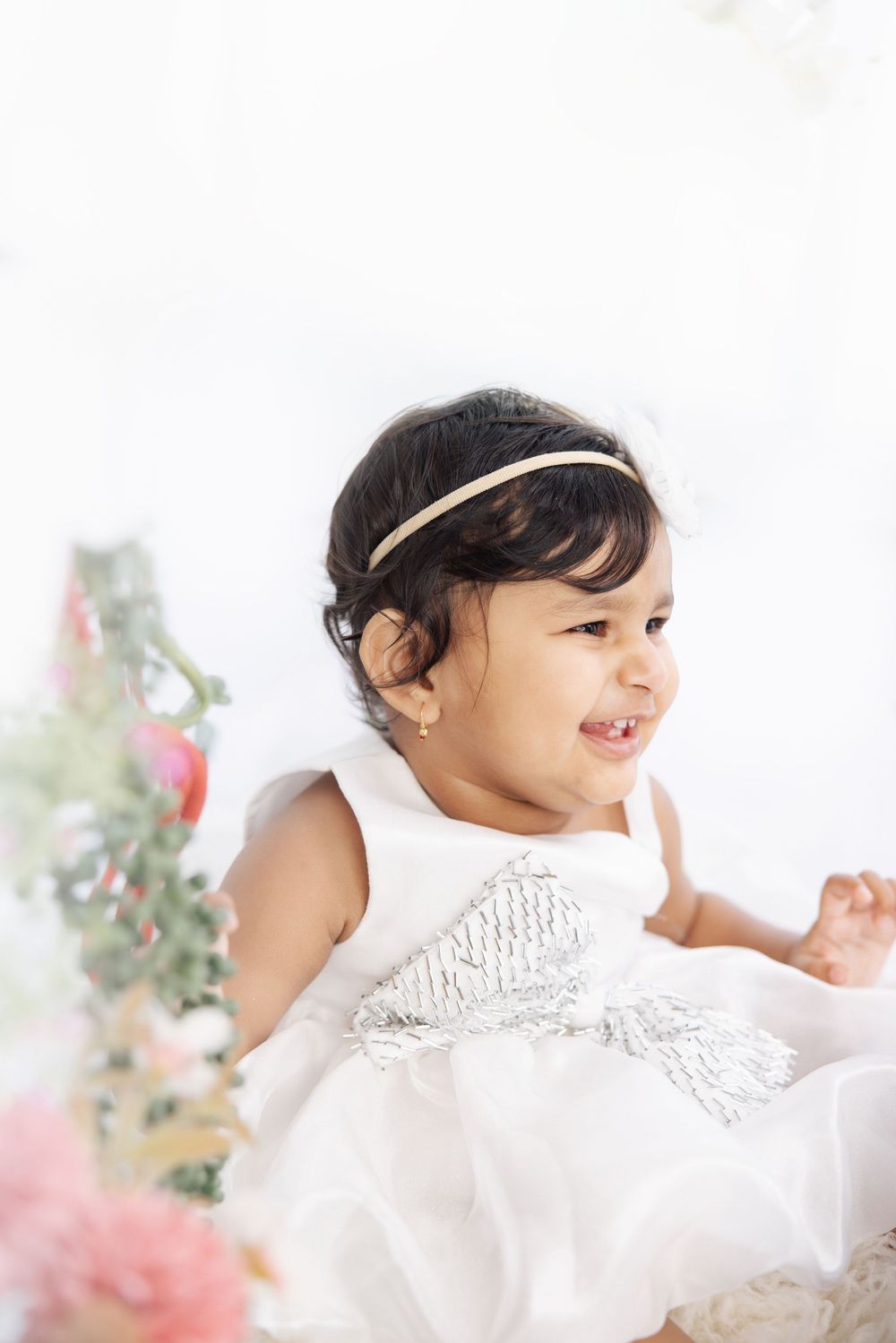 Smiling toddler girl in white sequin dress and gold headband surrounded by soft floral decor.