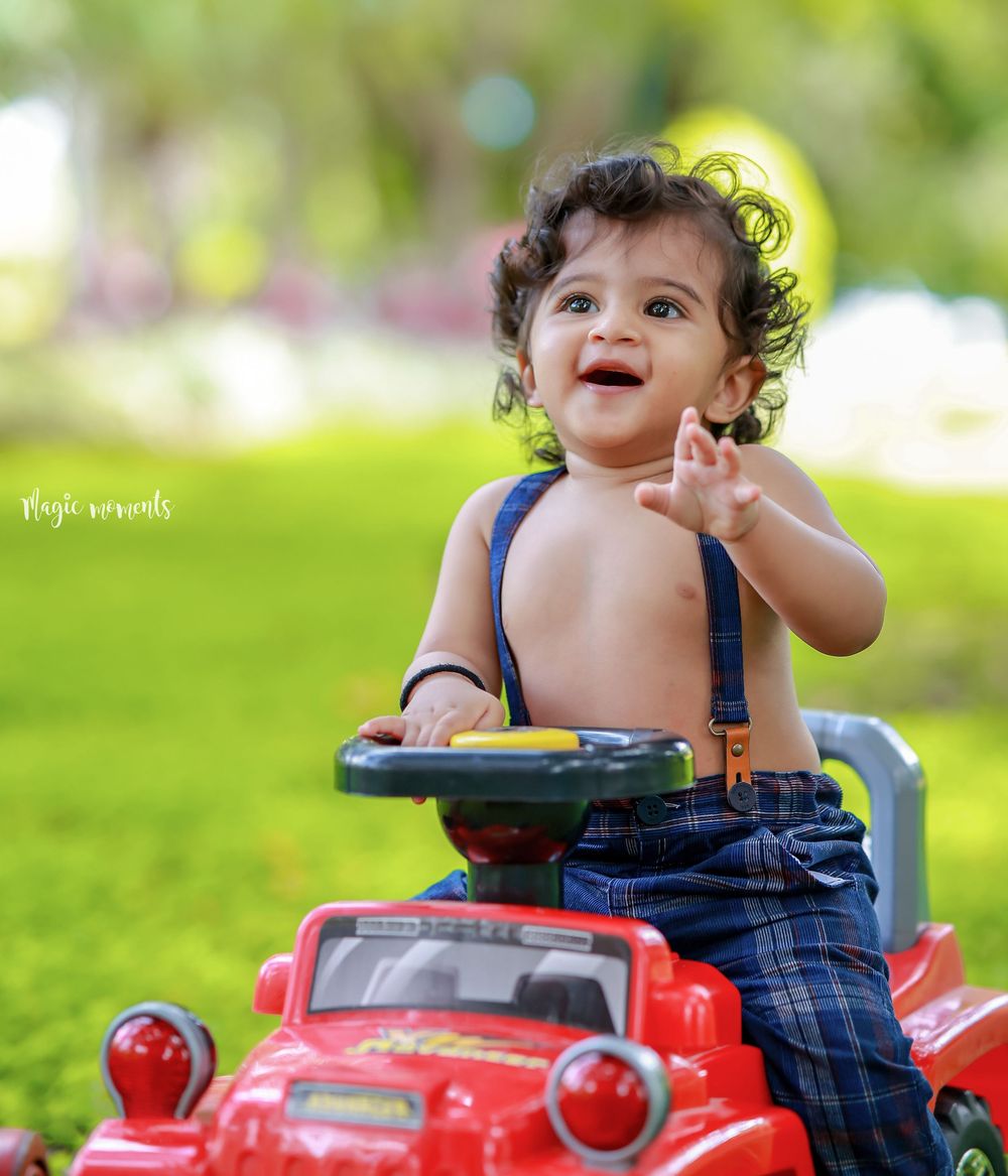 A happy toddler with curly hair sits in a red toy car wearing blue suspenders, smiling outdoors in a green park.