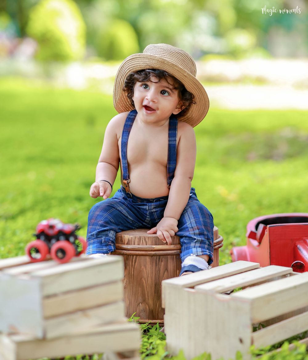 Cute toddler boy wearing straw hat and denim overalls sitting on wooden bucket outdoors in green park with toys.
