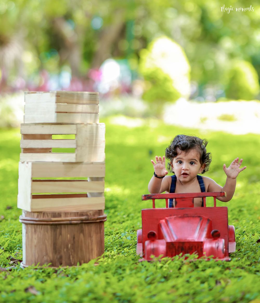 Toddler sitting in a red toy car on green grass, with stacked wooden crates beside them, hands raised in joy.