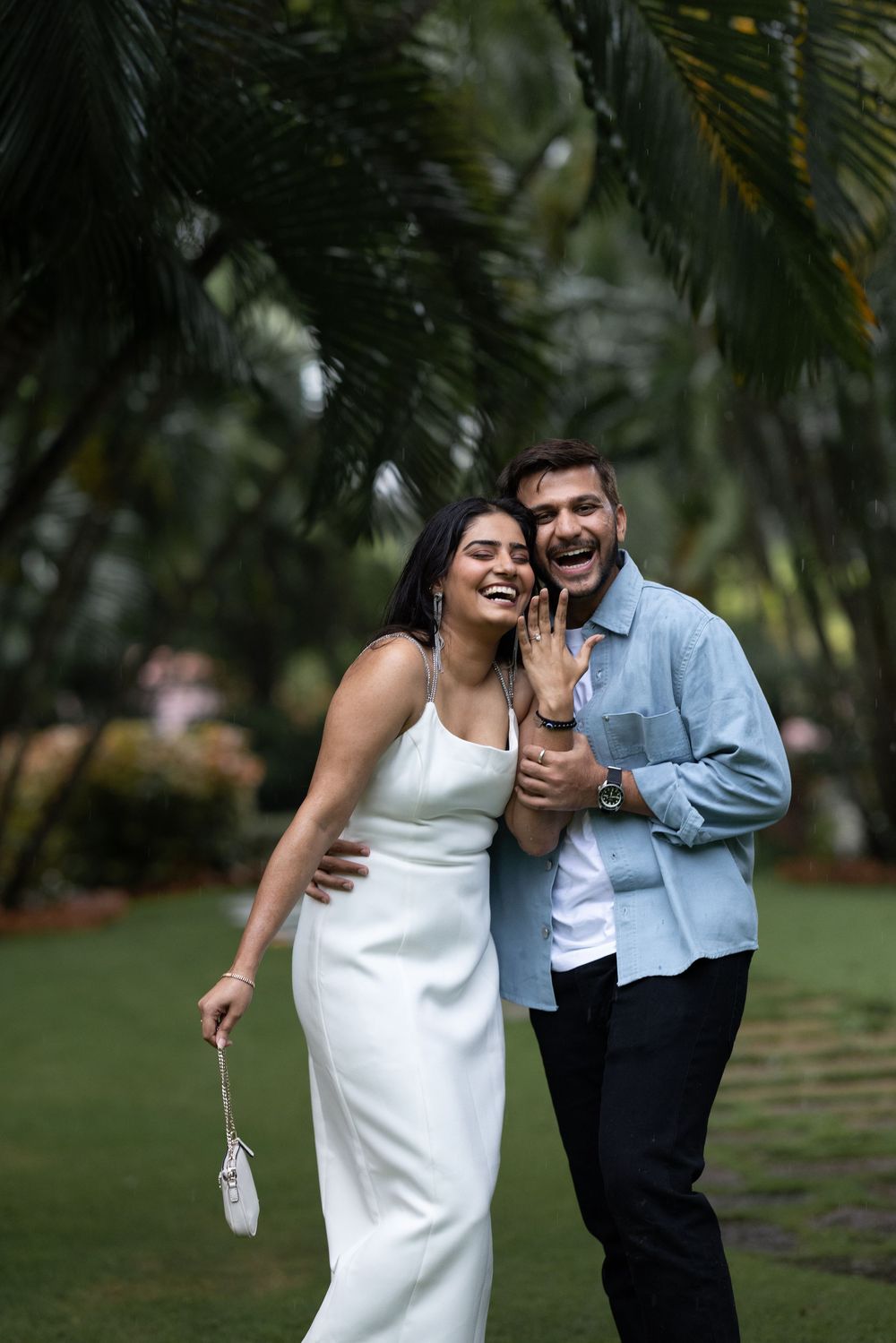 Joyful couple laughing outdoors, woman in white dress showing engagement ring, surrounded by tropical palm trees.