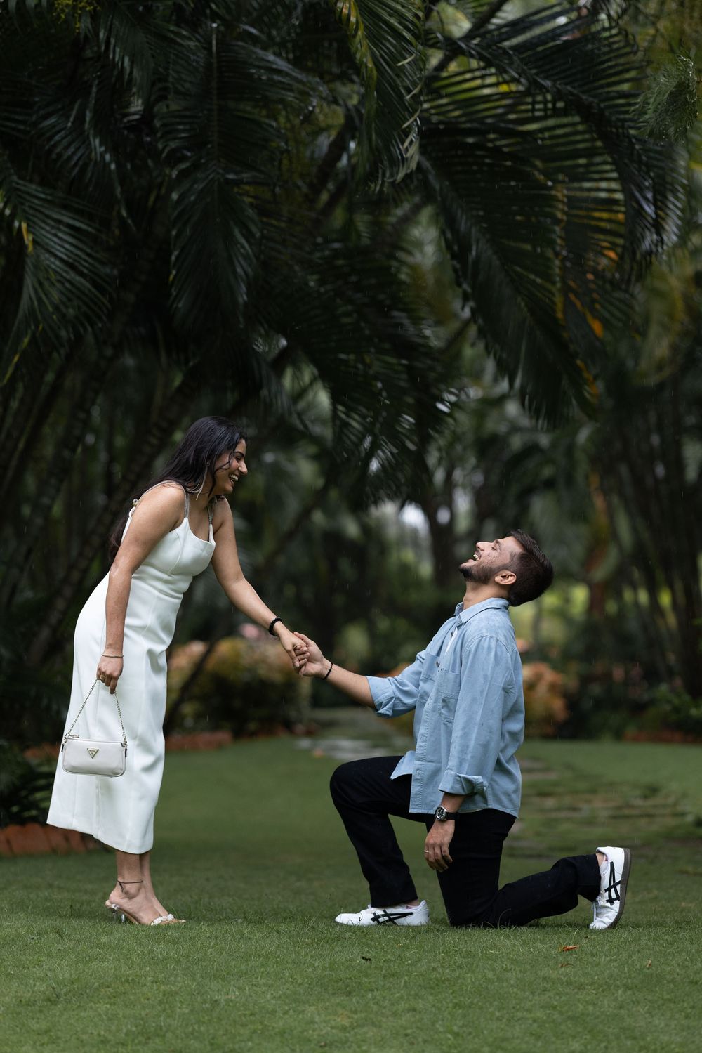 Man kneeling on one knee proposing to woman in white dress in a lush tropical garden setting.