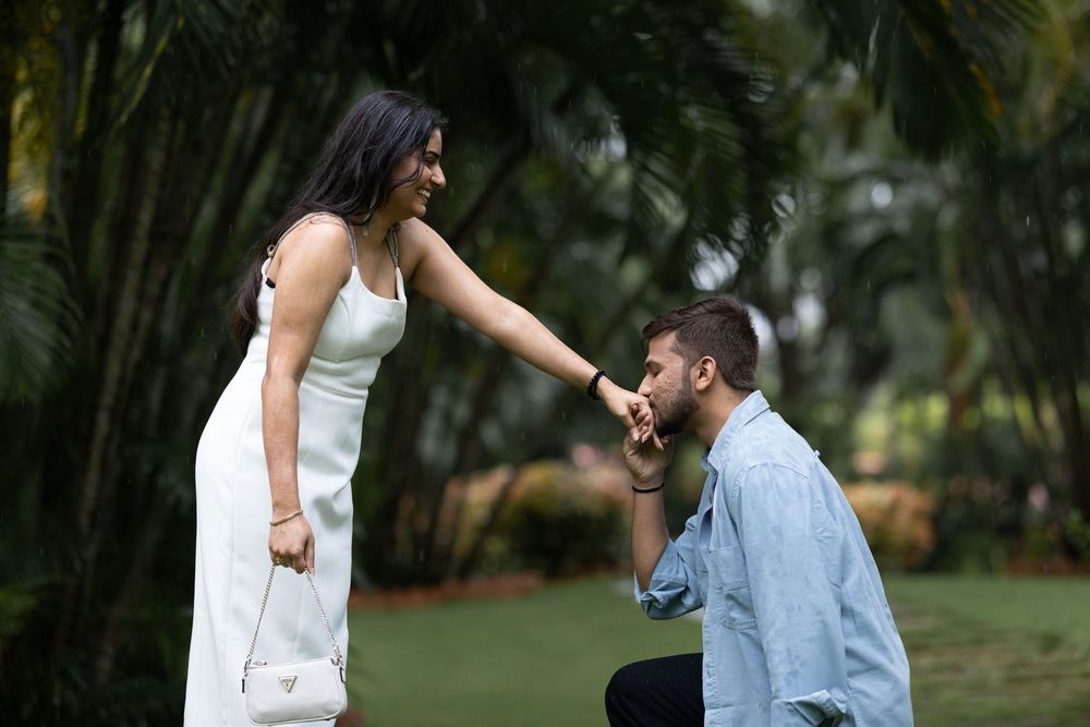 Romantic marriage proposal moment with man on one knee kissing woman's hand in a green tropical garden.
