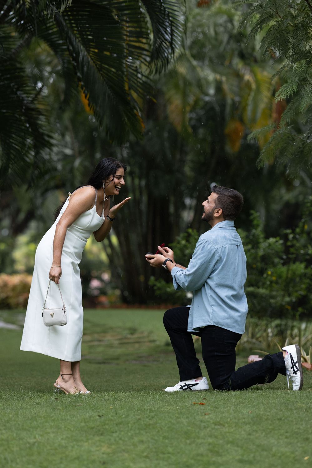 Excited woman reacting to surprise marriage proposal from man kneeling with ring box in tropical garden.
