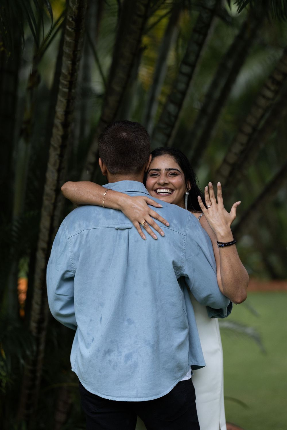 Newly engaged woman hugging her fiance while showing off diamond ring, smiling joyfully in tropical garden.