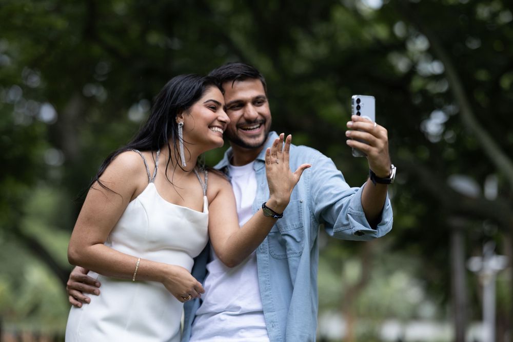 Smiling couple taking a selfie outdoors in park, woman in white dress, man in blue shirt, waving at camera.