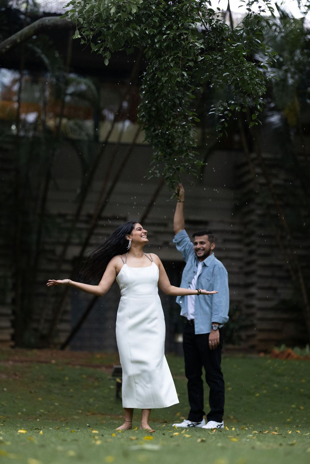 Man in denim shirt spinning woman in white dress under tree branches in garden, laughing together.
