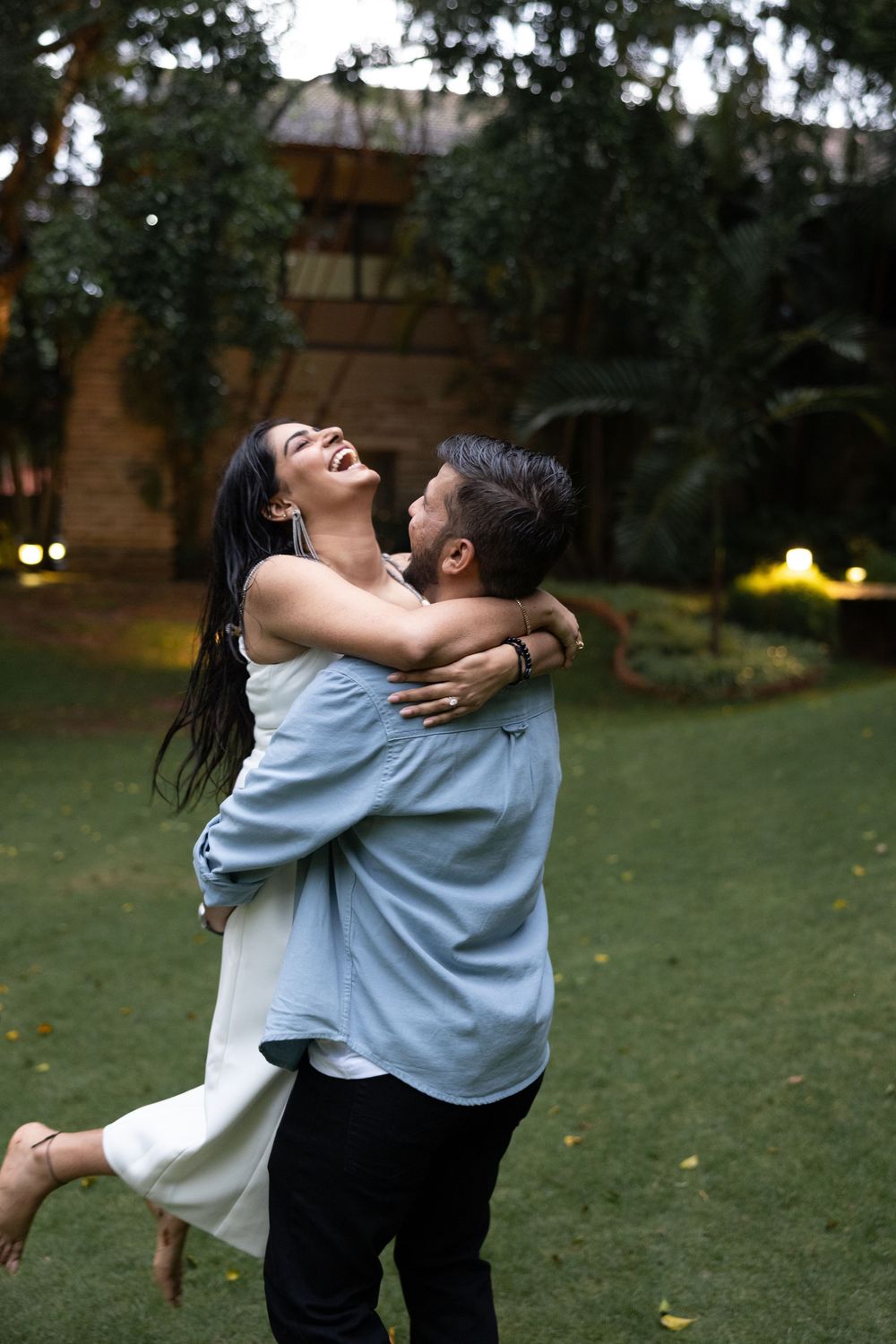 Man lifting and embracing laughing woman in white dress in garden at dusk, romantic couple moment.
