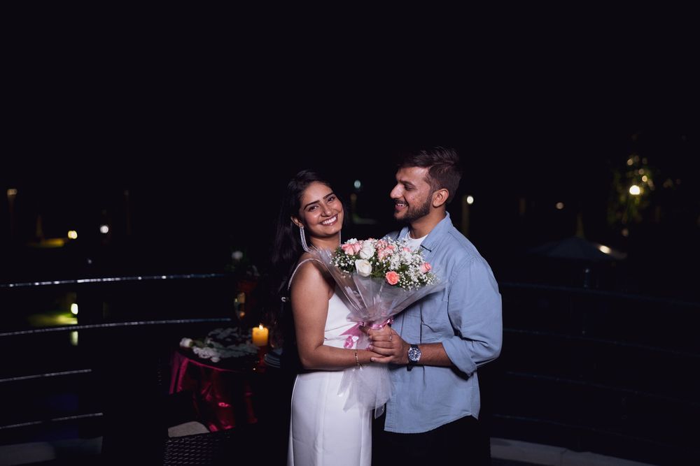 Couple smiling together at night holding pink flower bouquet, woman in white dress, man in blue shirt.