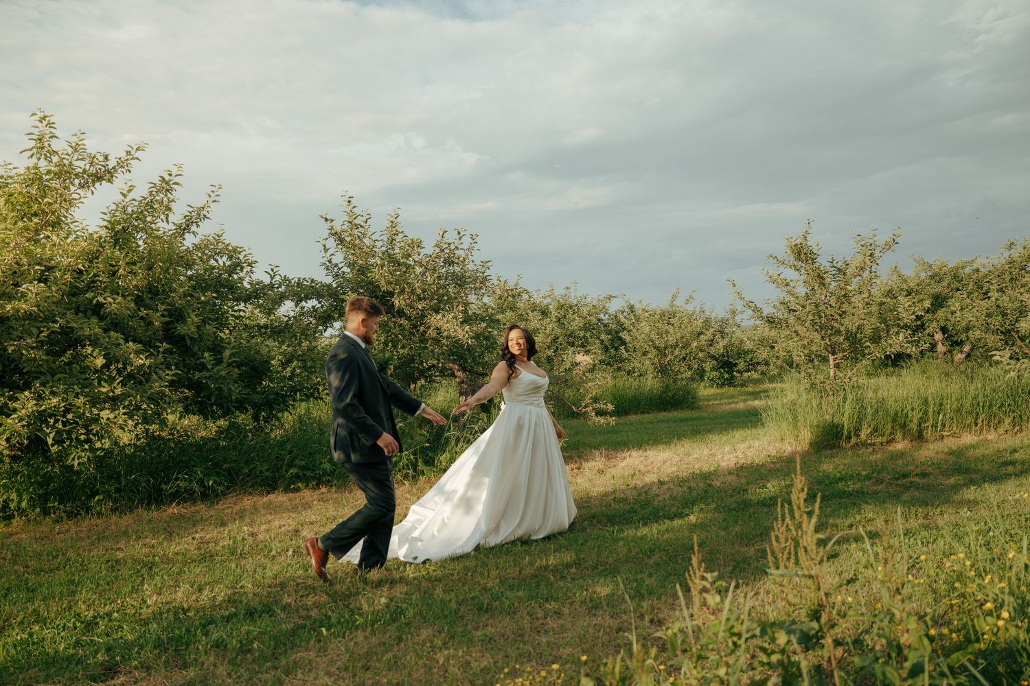 Bride in white gown and veil holds bouquet while groom in dark suit stands beneath green trees at outdoor wedding.