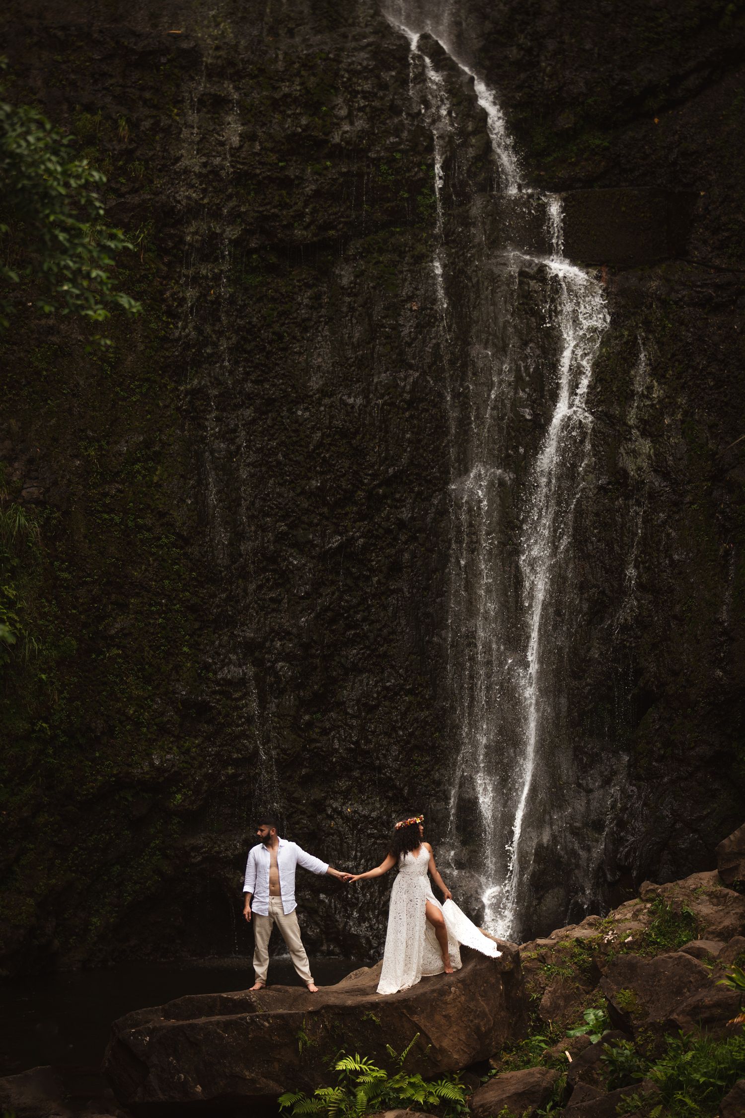 Couple holds hands on rocks before a dramatic cascading waterfall in a lush tropical setting.