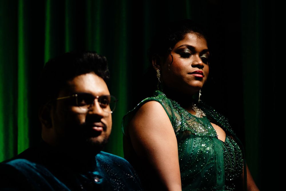 Two elegantly dressed South Asian individuals in green sequined outfits stand before a green curtain backdrop.