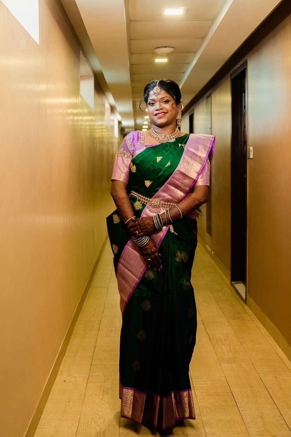 Indian bride in green and purple silk saree with gold jewelry and mehndi posing in hotel corridor.