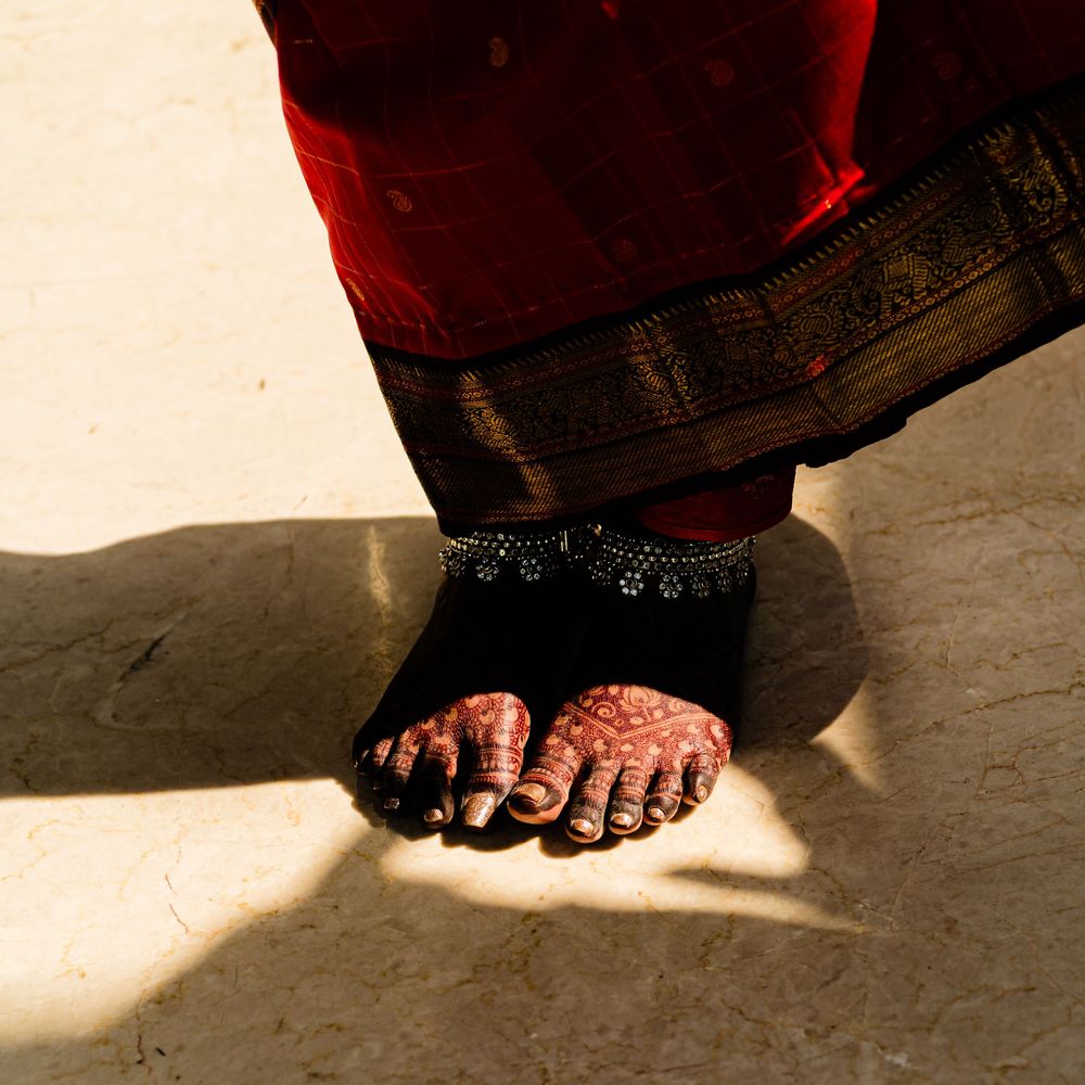 Close-up of feet adorned with henna and anklets, wearing a red and gold sari, standing on a sunlit floor.
