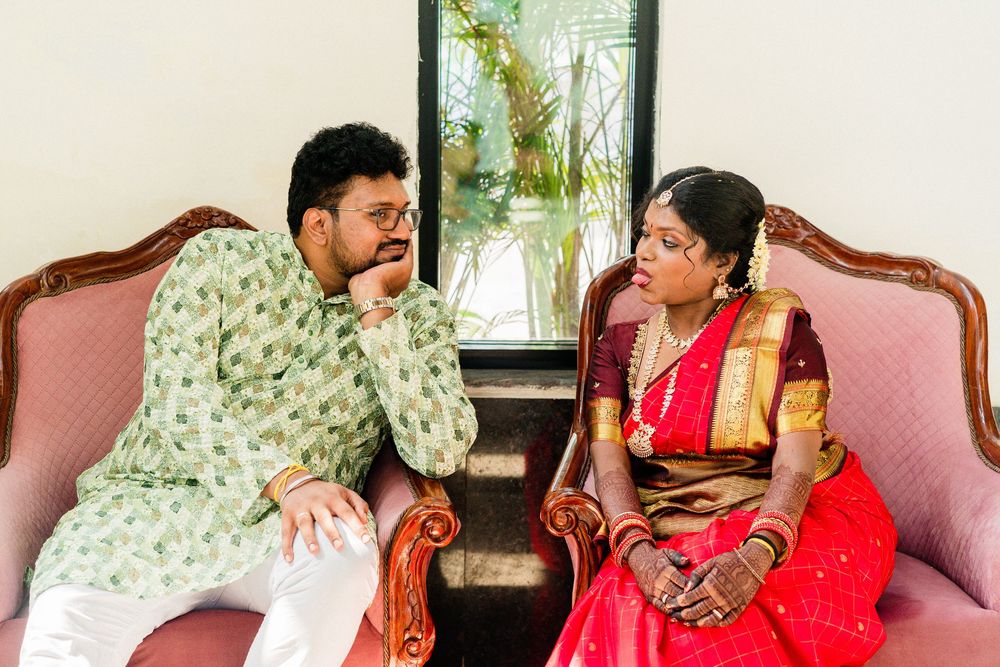 Indian couple in traditional wedding attire seated on pink sofa, bride in red saree, groom in green kurta.