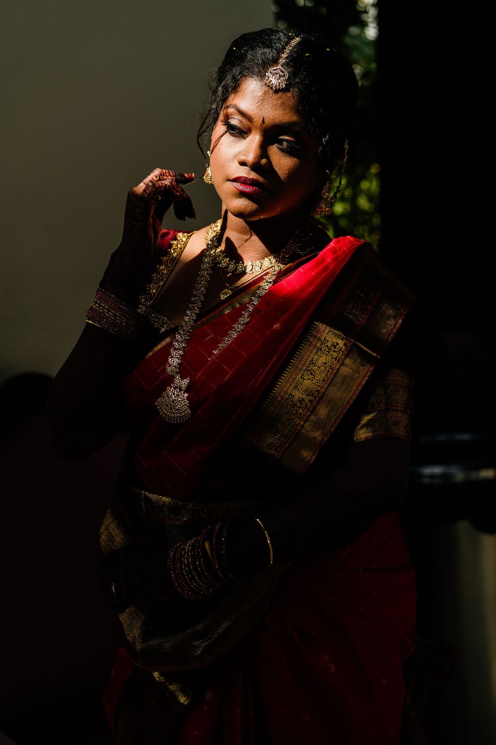 Indian bride in red and gold saree with traditional jewelry poses in dramatic lighting.