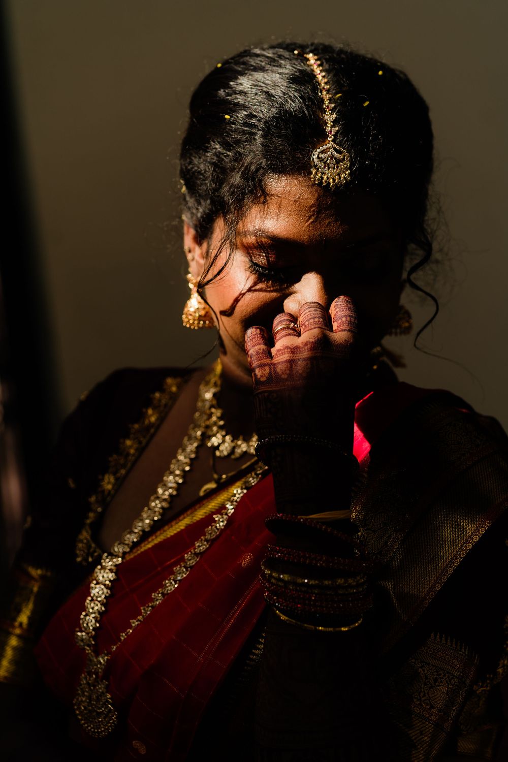 Indian bride in red and gold saree with traditional jewelry covering her face emotionally.