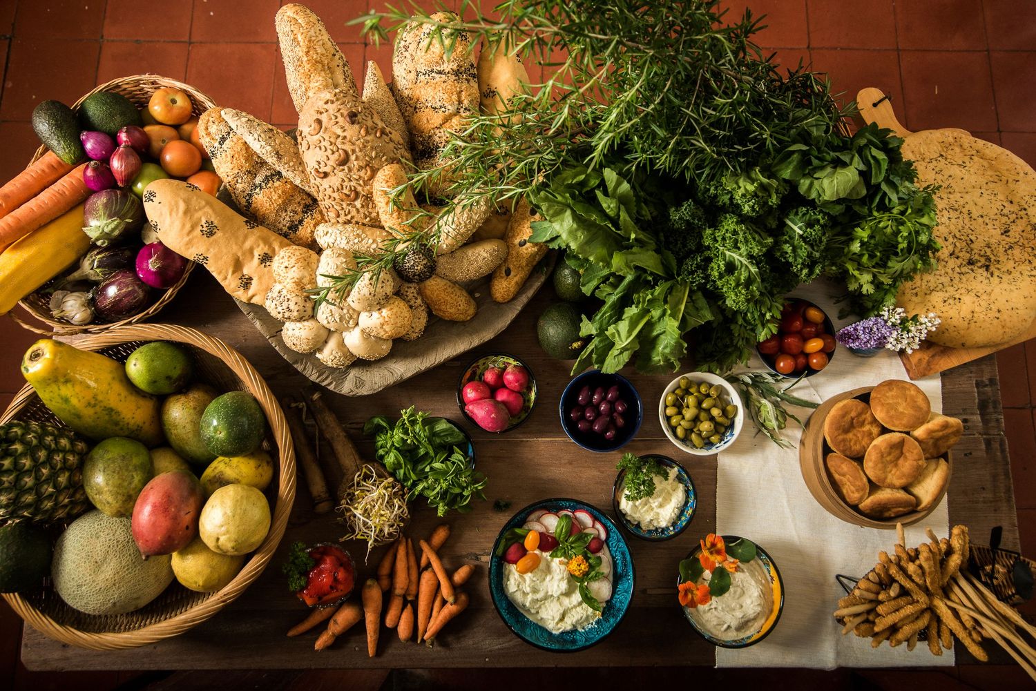 Overhead view of a rustic wooden table filled with fresh vegetables, fruits, herbs, breads, and dips in a vibrant spread.