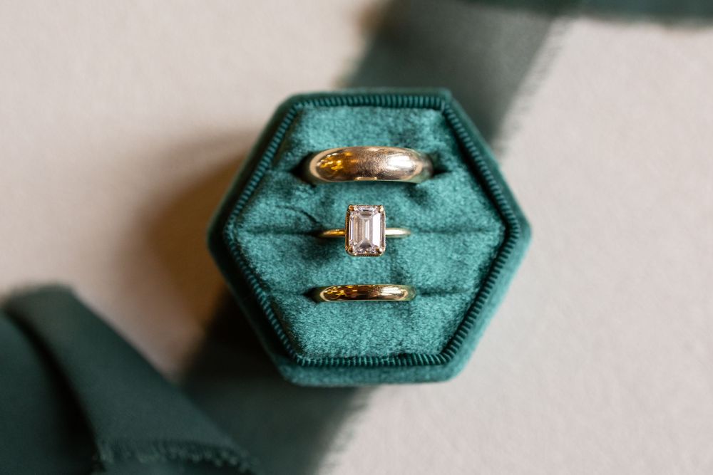 Three gold rings displayed in a teal hexagonal velvet ring box, featuring an emerald cut diamond engagement ring.