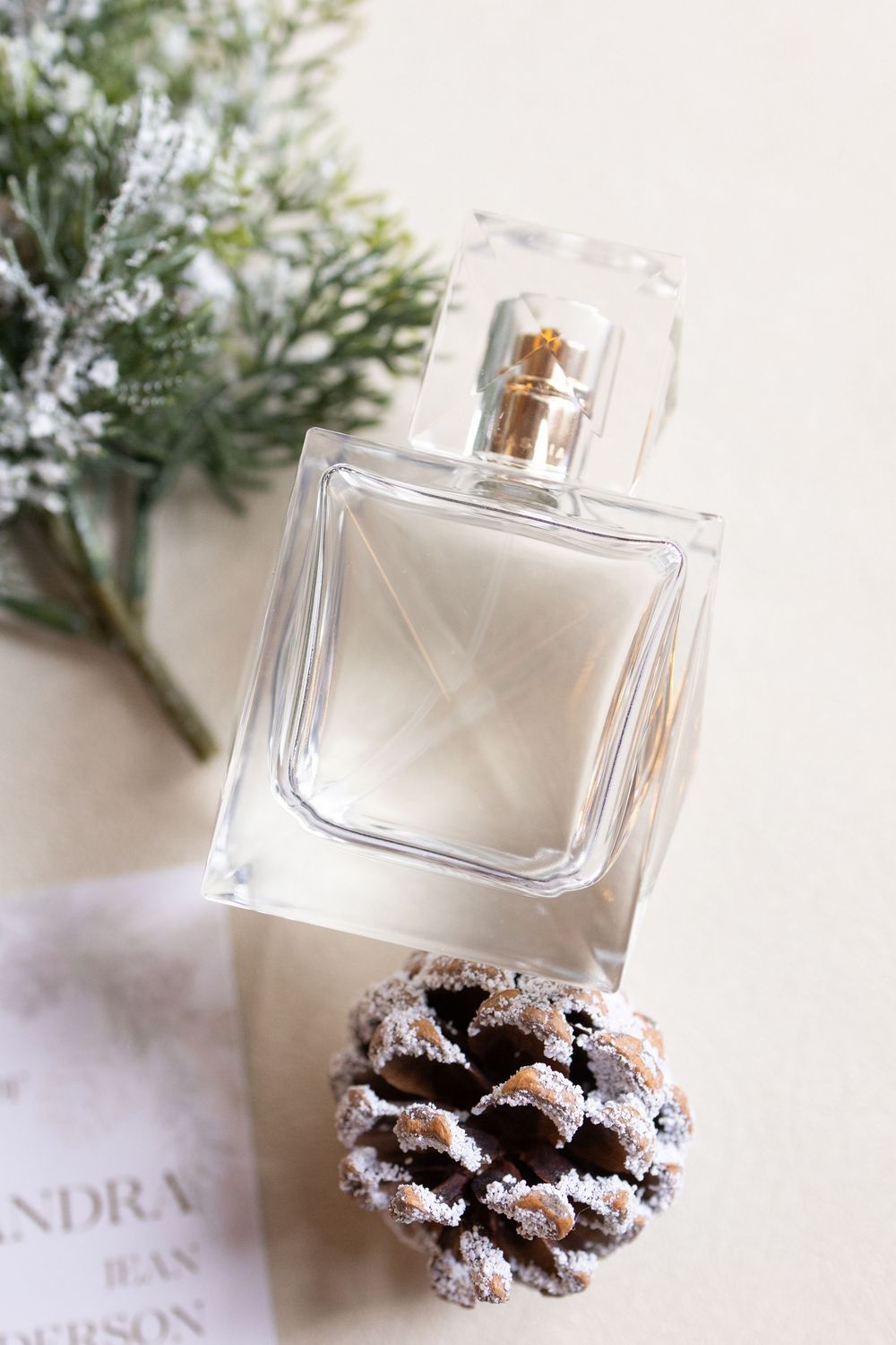 Clear glass perfume bottle with pine cone and frosted evergreen branches on white background, winter holiday styling.