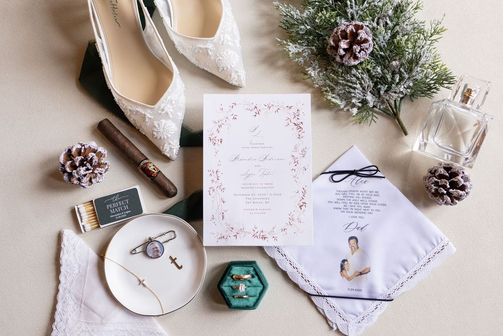 Elegant wedding flat lay with lace heels, floral invitation, rings, pine cones, perfume, and bridal accessories on neutral background.
