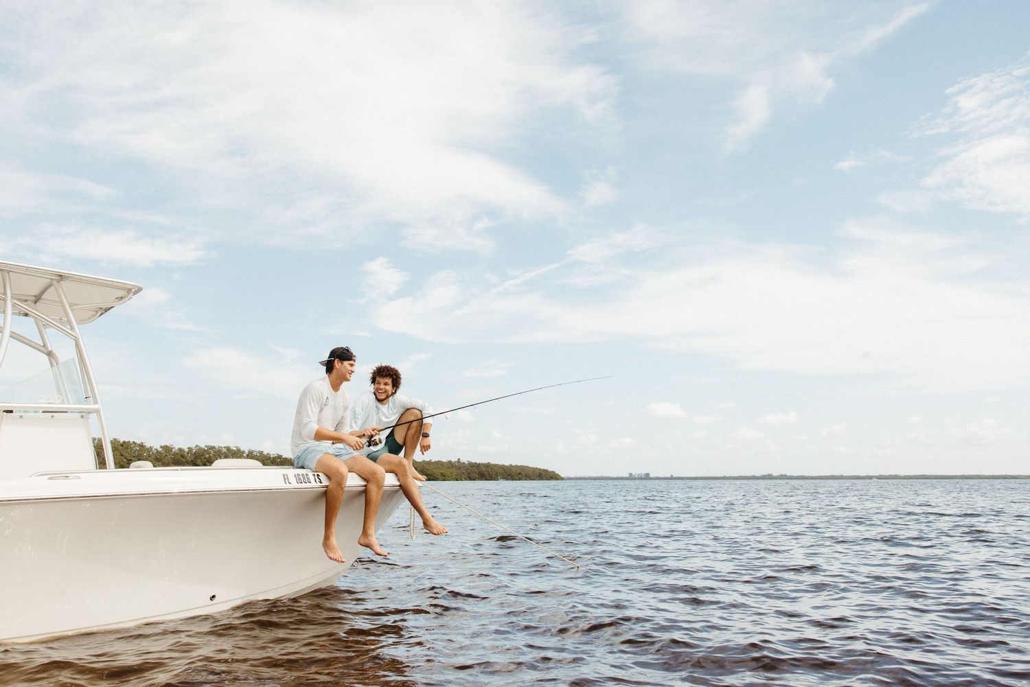 Two people sitting on the bow of a boat fishing over calm water with a tree-lined horizon under a cloudy sky.