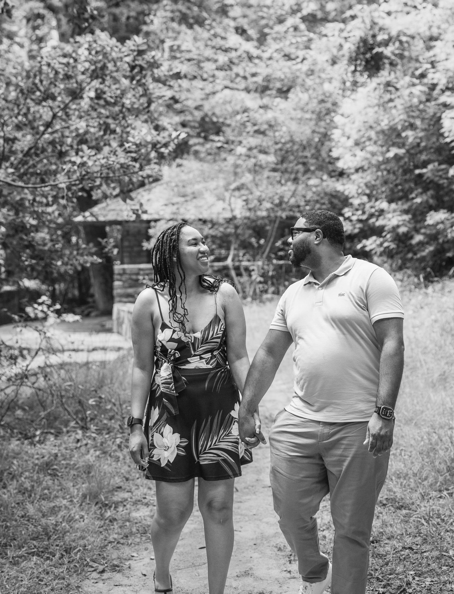 Black and white photo of a smiling couple holding hands while walking on a dirt path surrounded by lush trees.