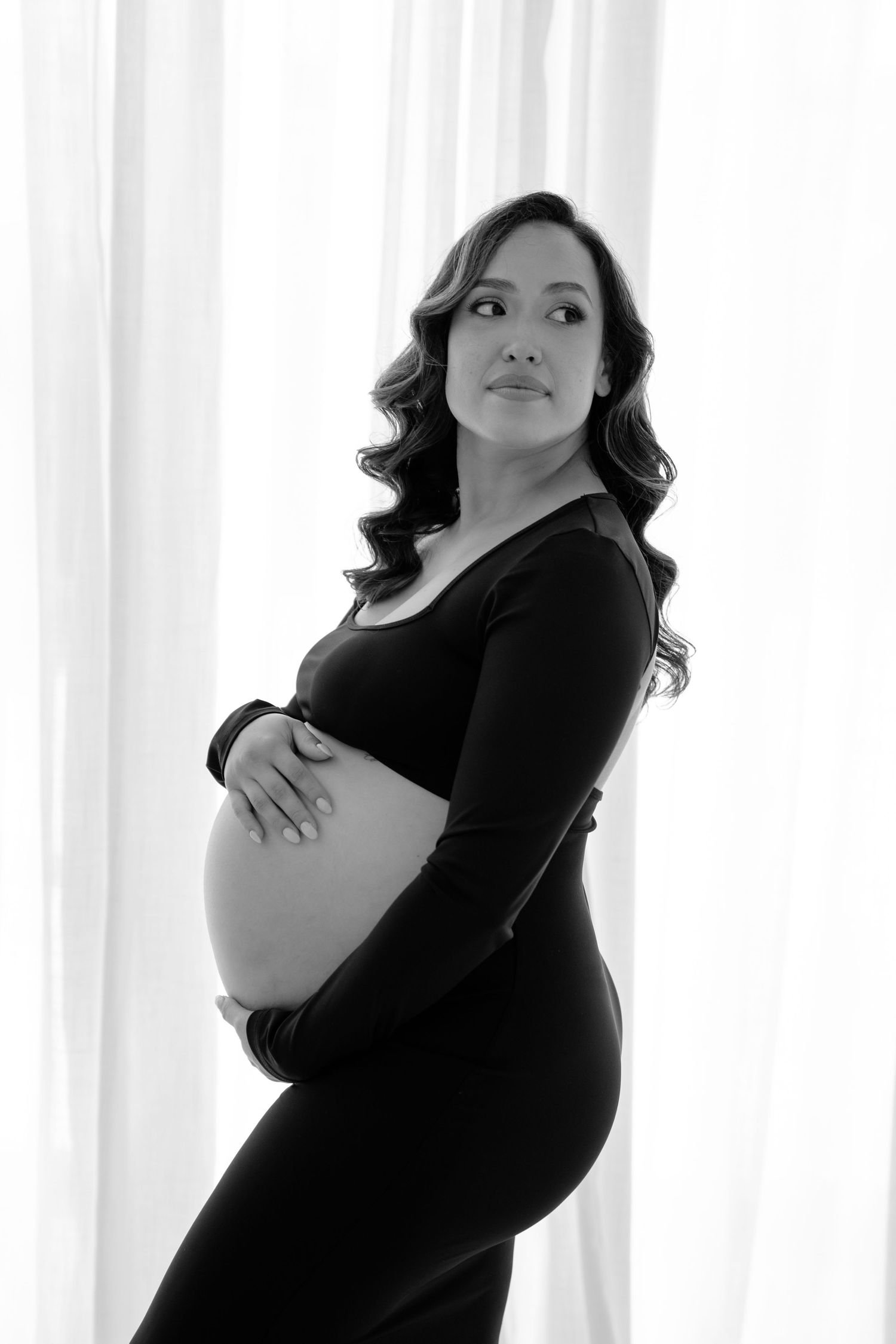 Black and white portrait of a pregnant woman in a black dress cradling her baby bump near sheer white curtains.