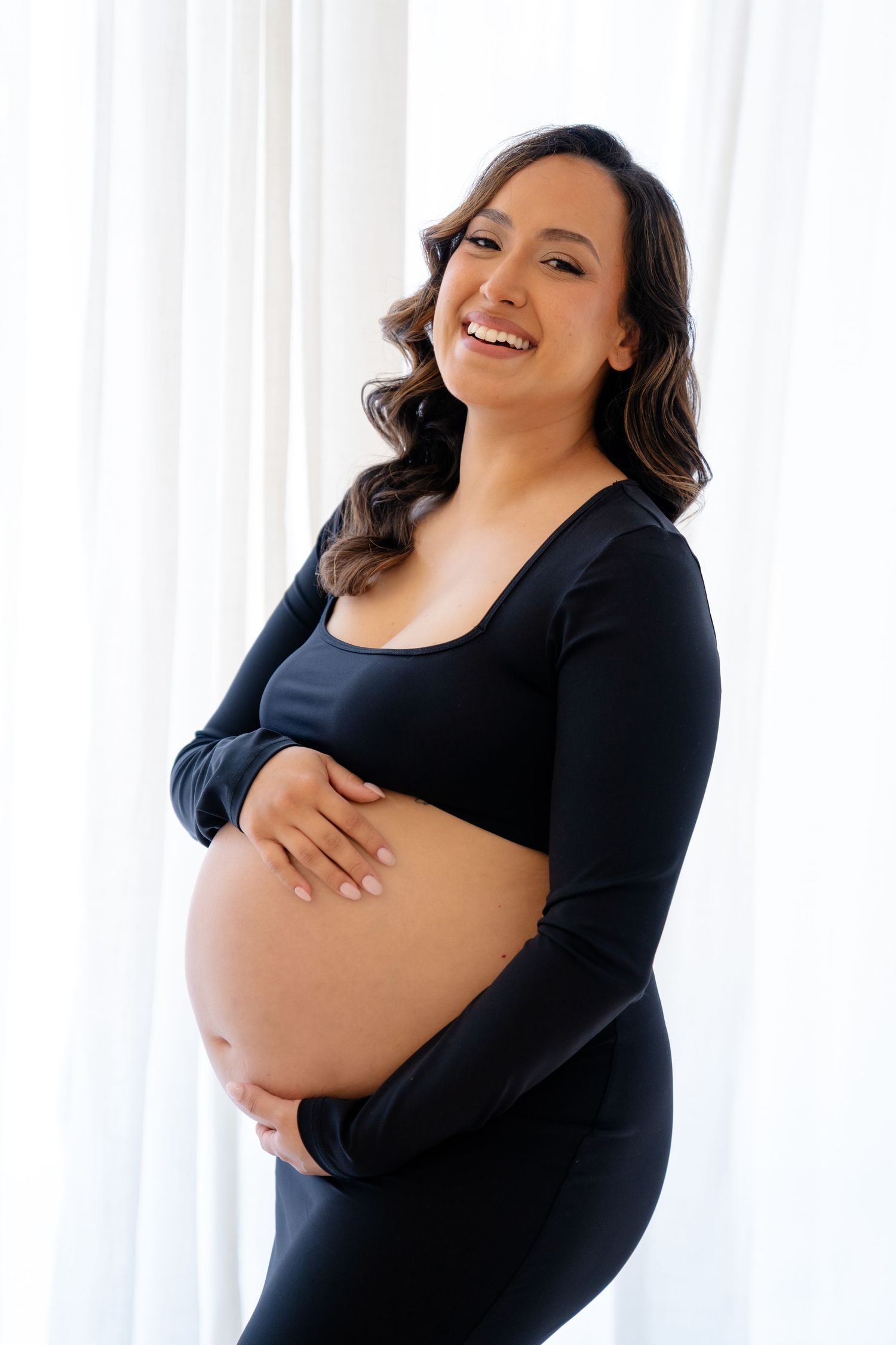Smiling pregnant woman in black outfit cradling baby bump against white curtain backdrop.