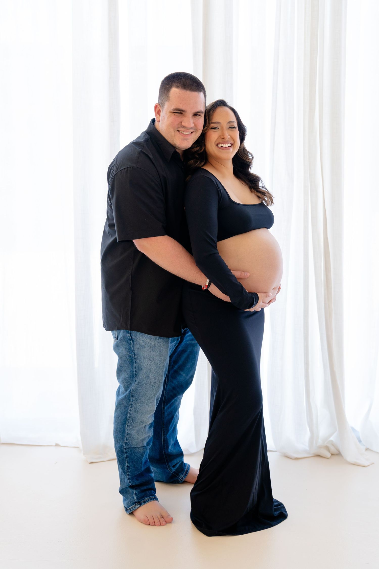 Smiling couple in maternity photo shoot, man in black shirt and jeans embracing pregnant woman in navy blue gown.