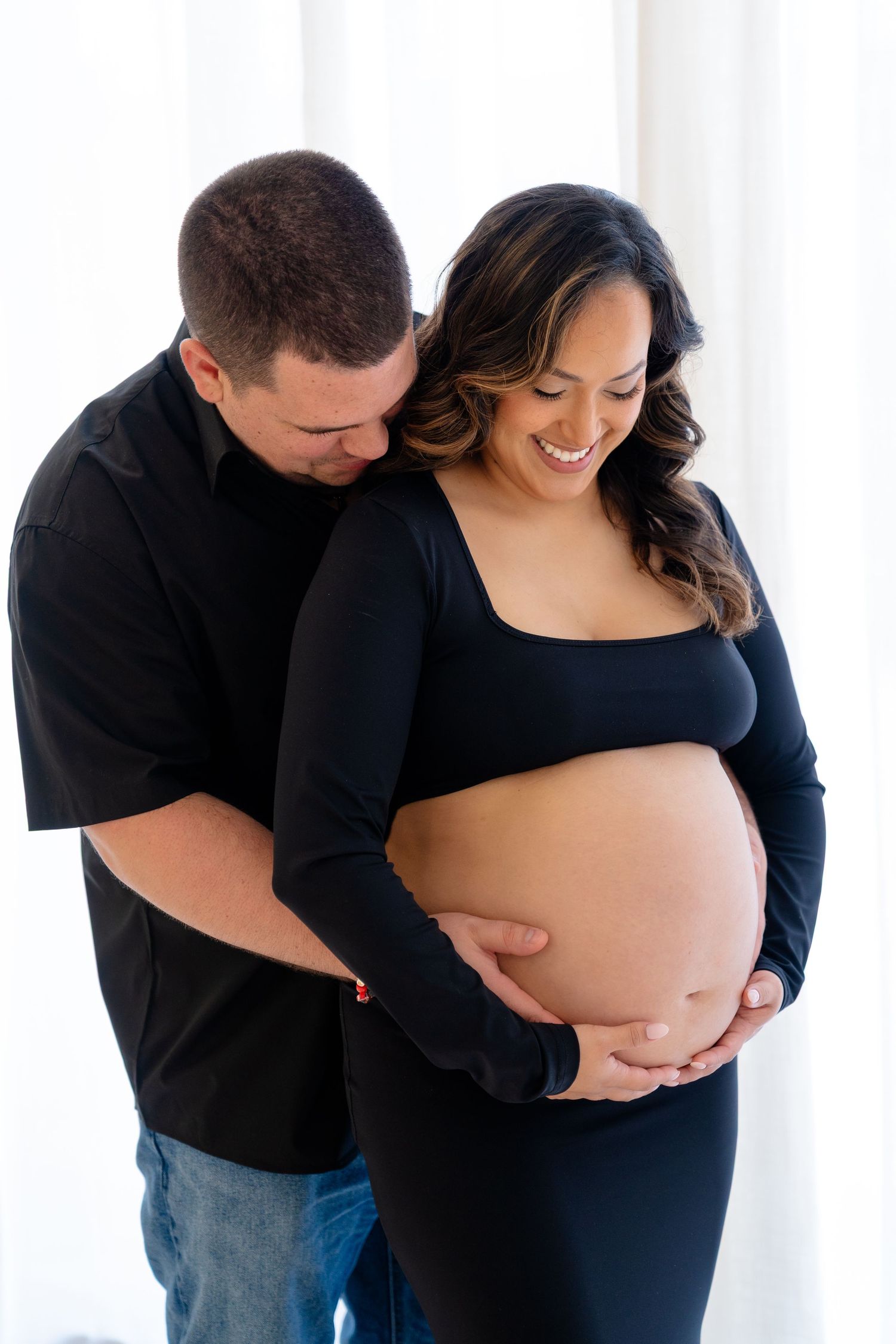Smiling pregnant woman in black outfit cradling baby bump while partner embraces her from behind near white curtains.