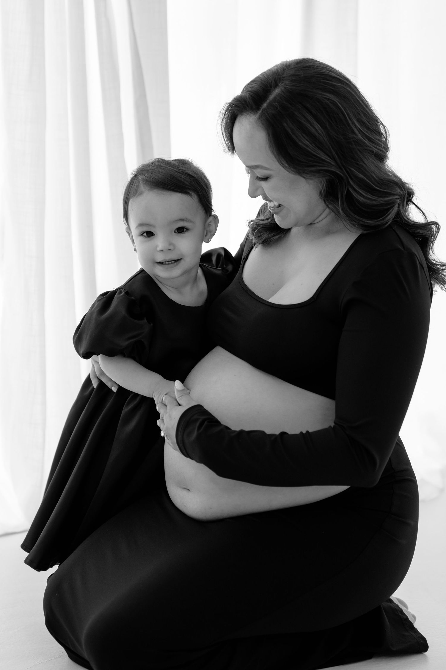 Black and white maternity photo of pregnant woman in black dress sitting with toddler daughter, both smiling warmly.