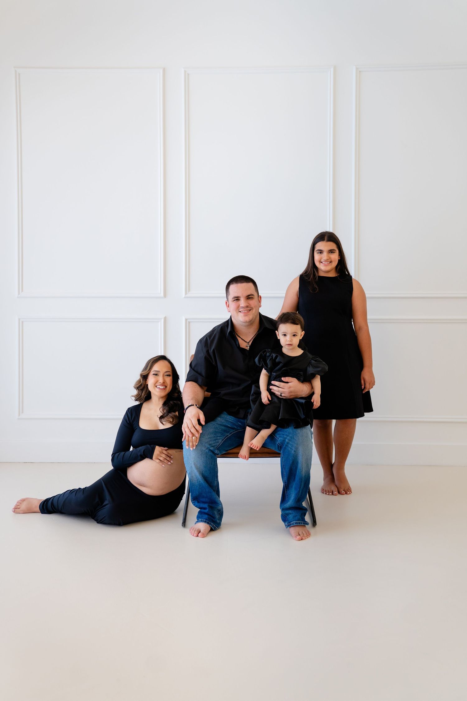 Family maternity photo with pregnant mom, dad, toddler, and teen in black outfits on white background.