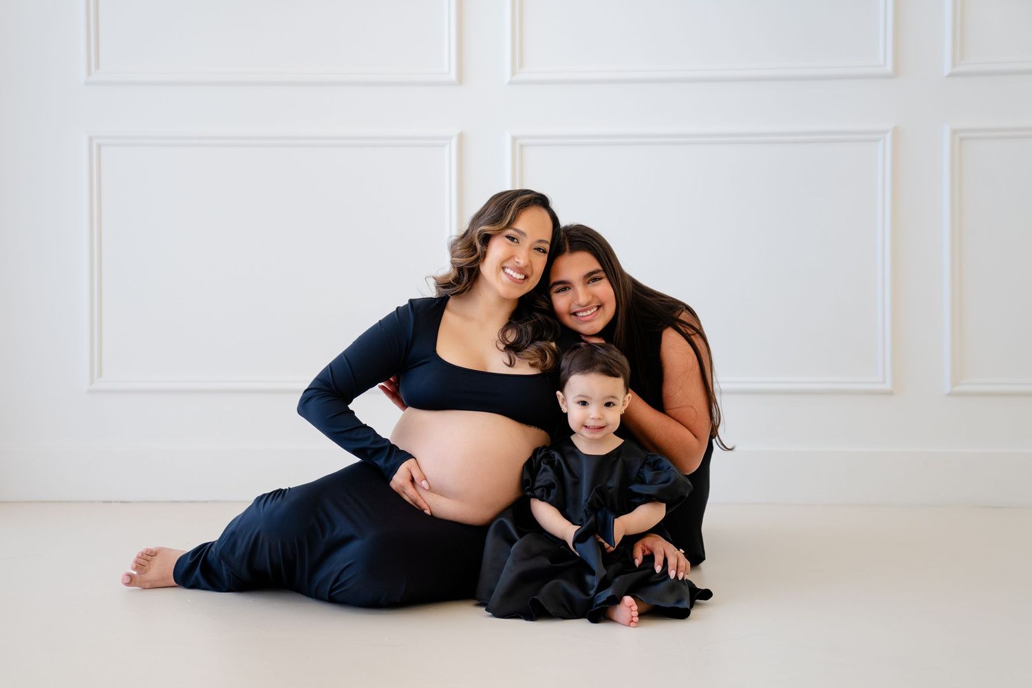 Pregnant woman in navy blue outfit poses with smiling woman and toddler in black against white studio backdrop.