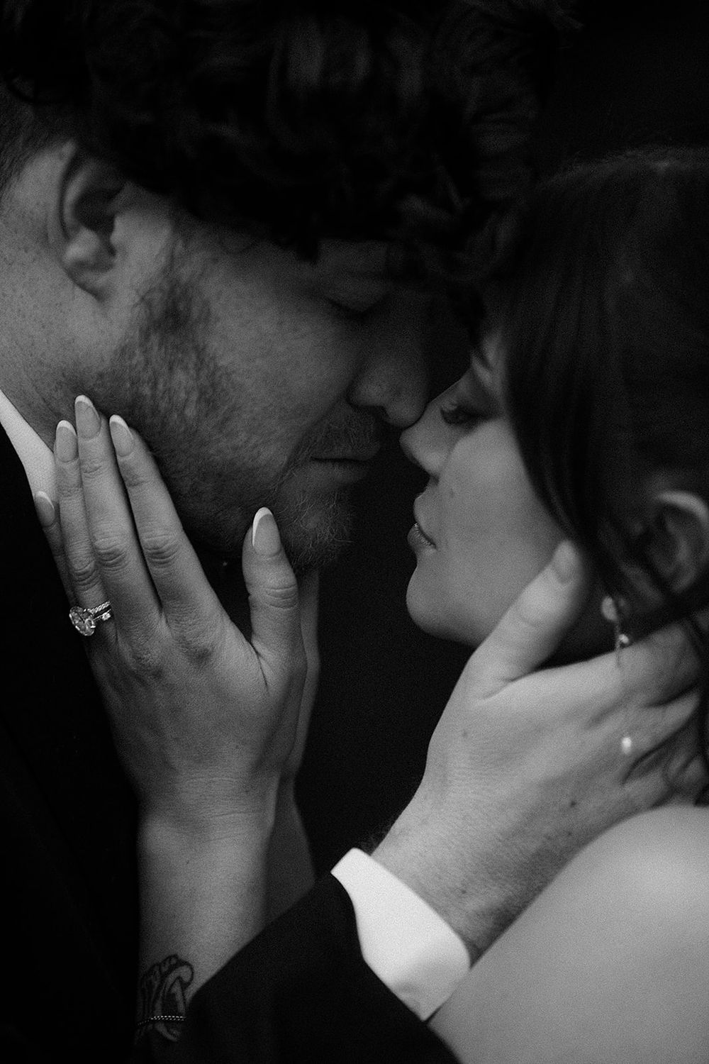Black and white photo of a couple sharing an intimate moment, the woman wearing an engagement ring as they lean close together.