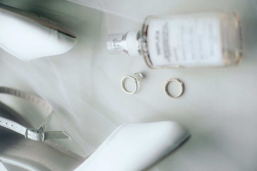 Two wedding rings rest beside a perfume bottle and white bridal heels on a light surface.