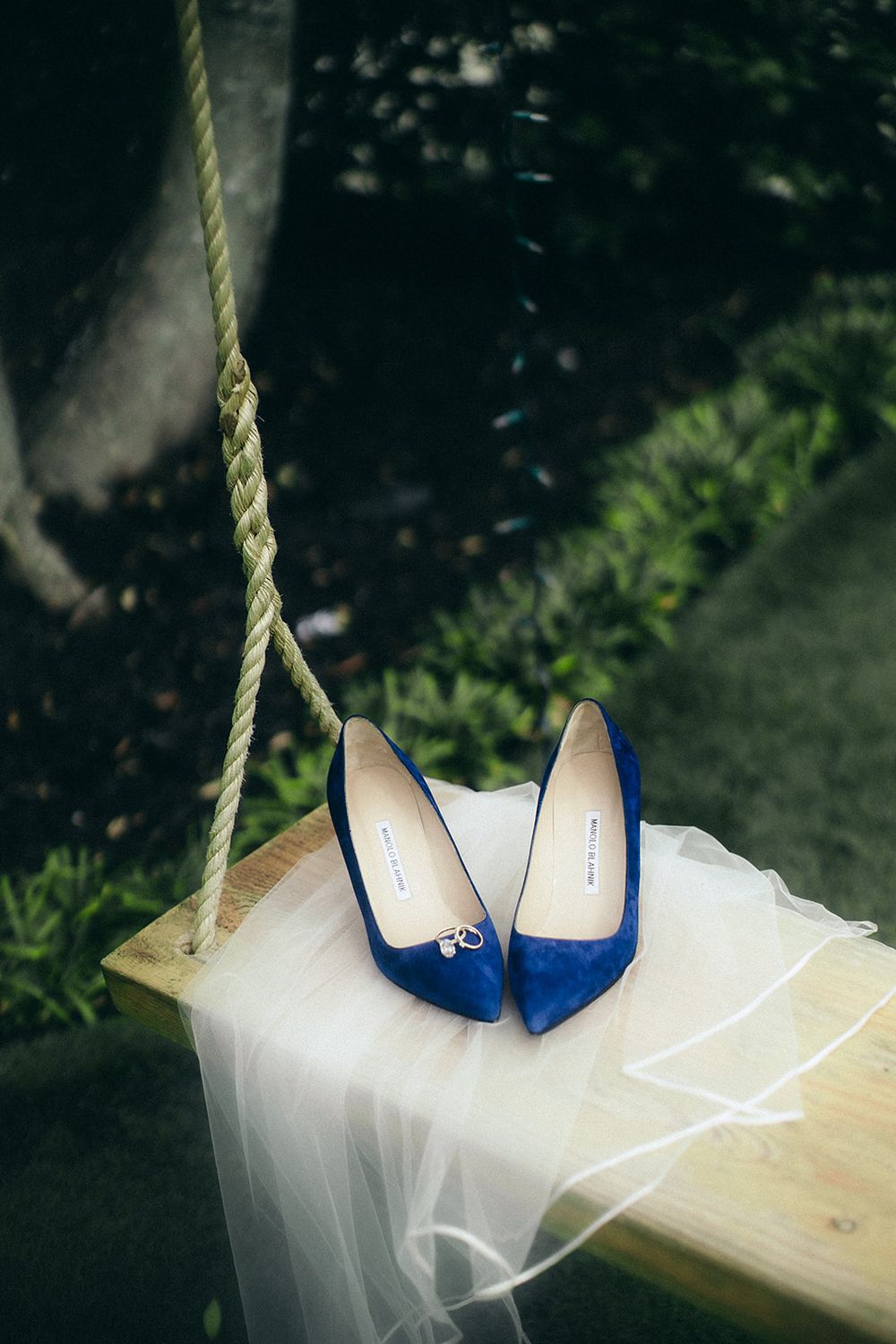Blue suede pointed-toe heels with bow detail resting on a wooden swing with white tulle veil outdoors.