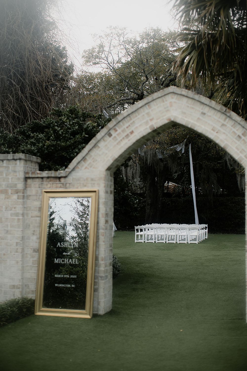 Elegant outdoor wedding ceremony setup with white chairs under a stone arch, featuring a gold-framed welcome mirror sign.