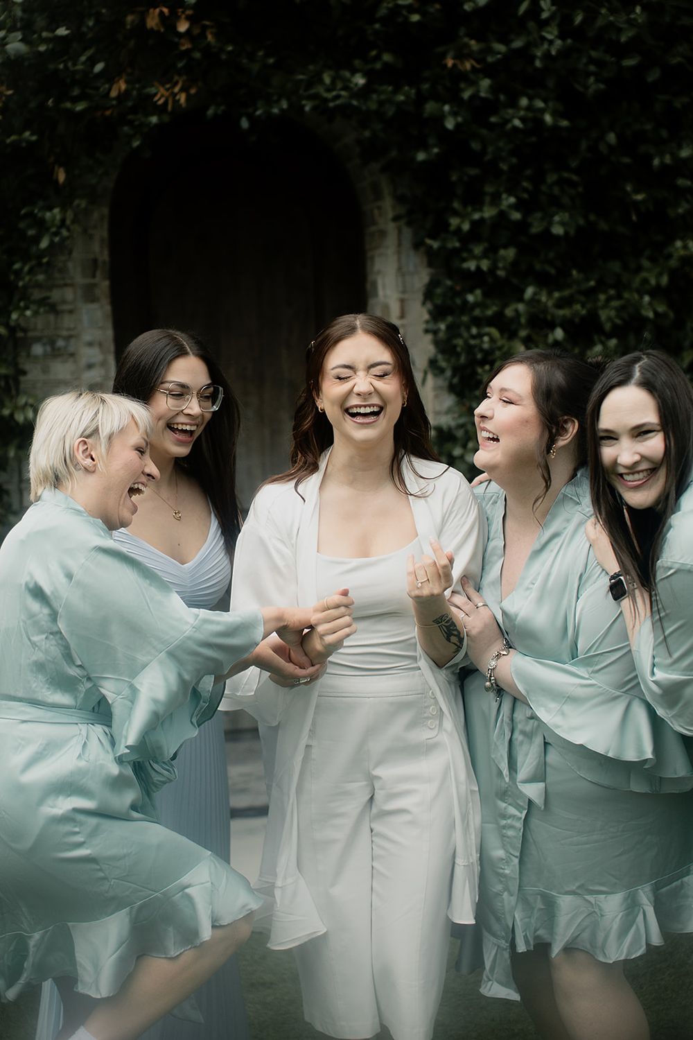 Bride in white robe laughing with four bridesmaids in mint satin robes outdoors near ivy-covered stone wall.
