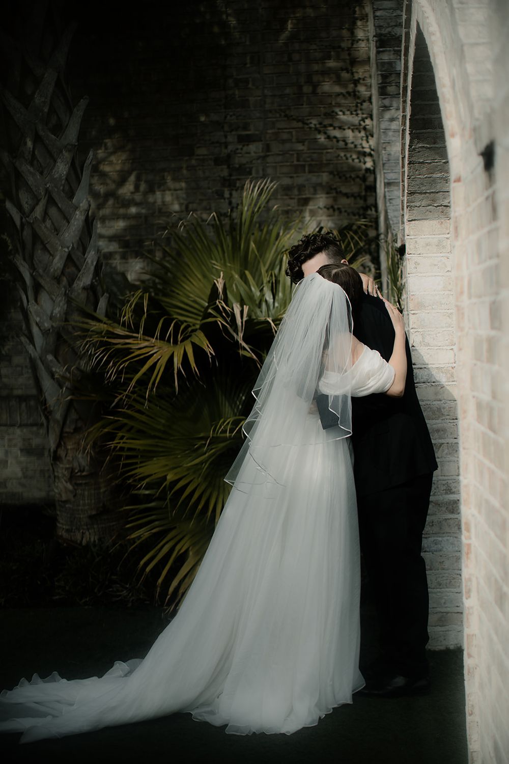 Bride in white gown and veil kissing groom in black suit against brick wall with tropical palms.