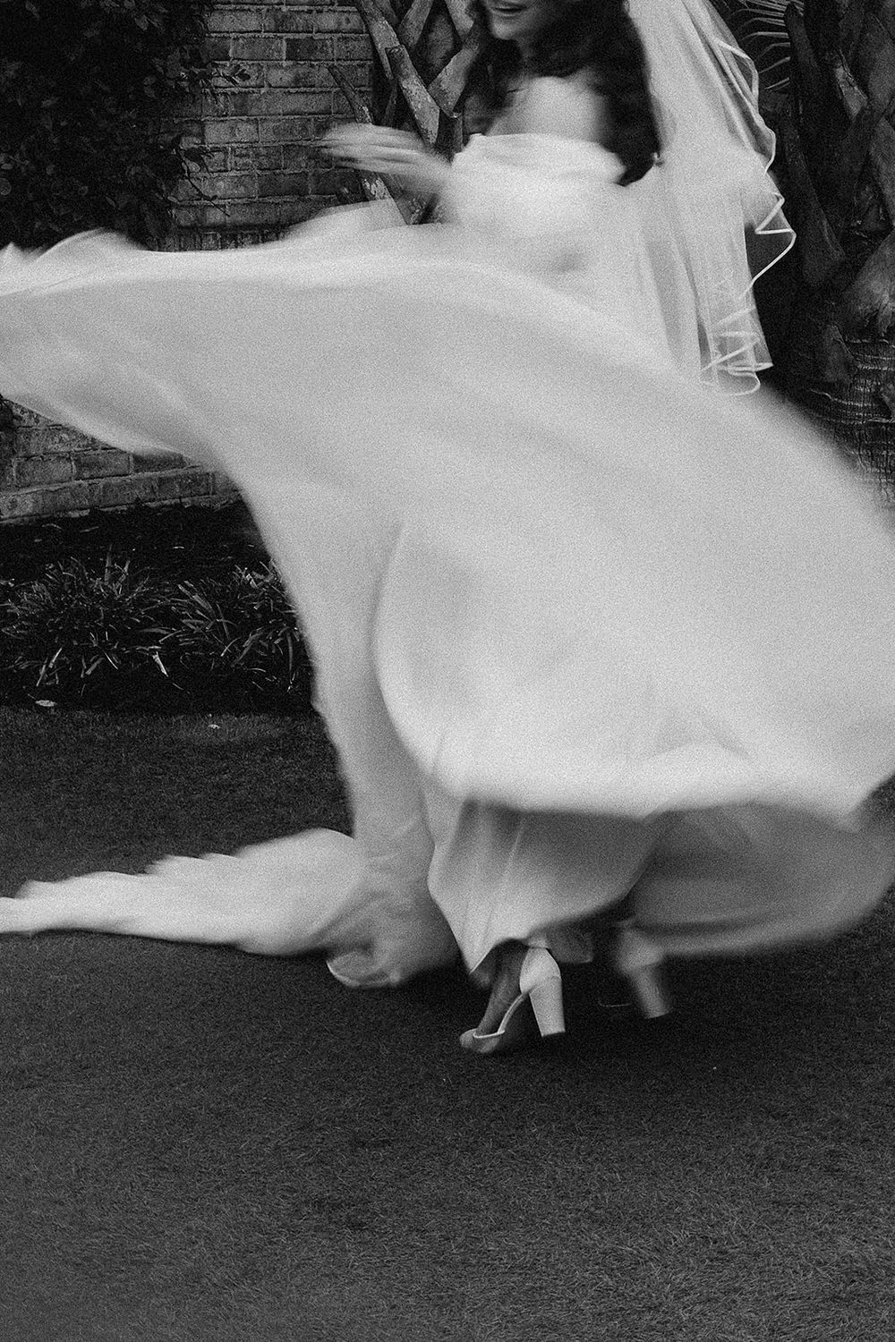 Black and white photo of a bride twirling in a flowing white wedding gown and veil outdoors.