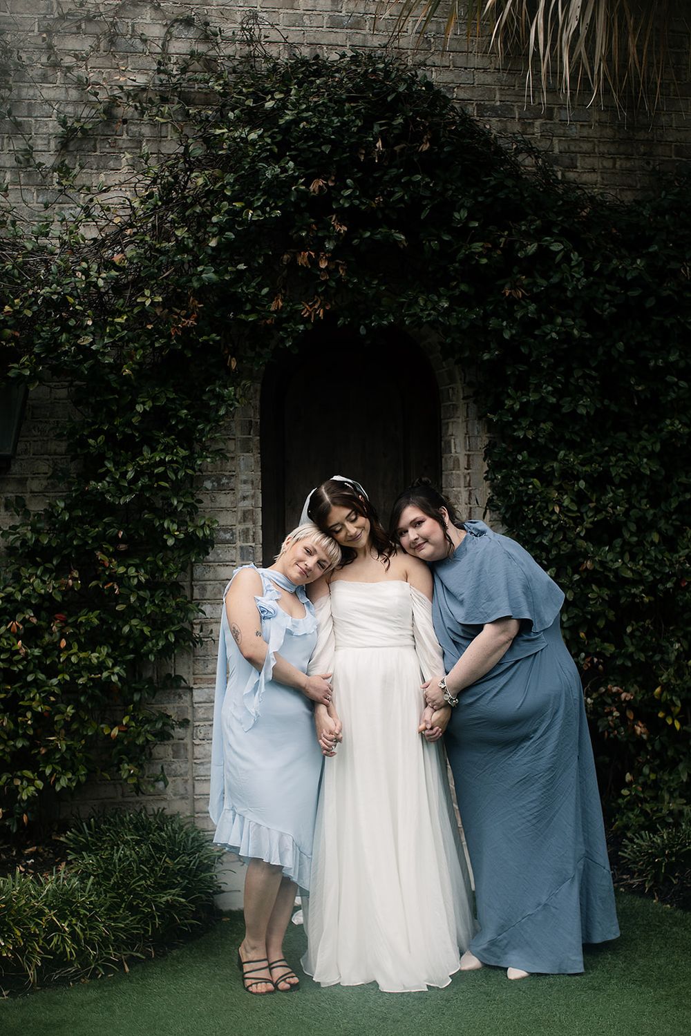 Bride in white gown poses with two bridesmaids in blue dresses before ivy-covered stone wall and wooden door.