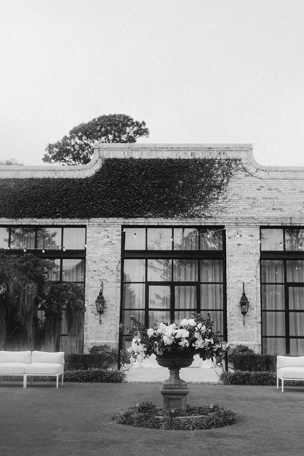 Black and white photo of an elegant brick venue with ivy, large windows, floral urn centerpiece, and white lounge seating on manicured lawn.